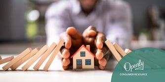 A toy wooden house is held between two hands, while dominos fall against the outside of both hands. 