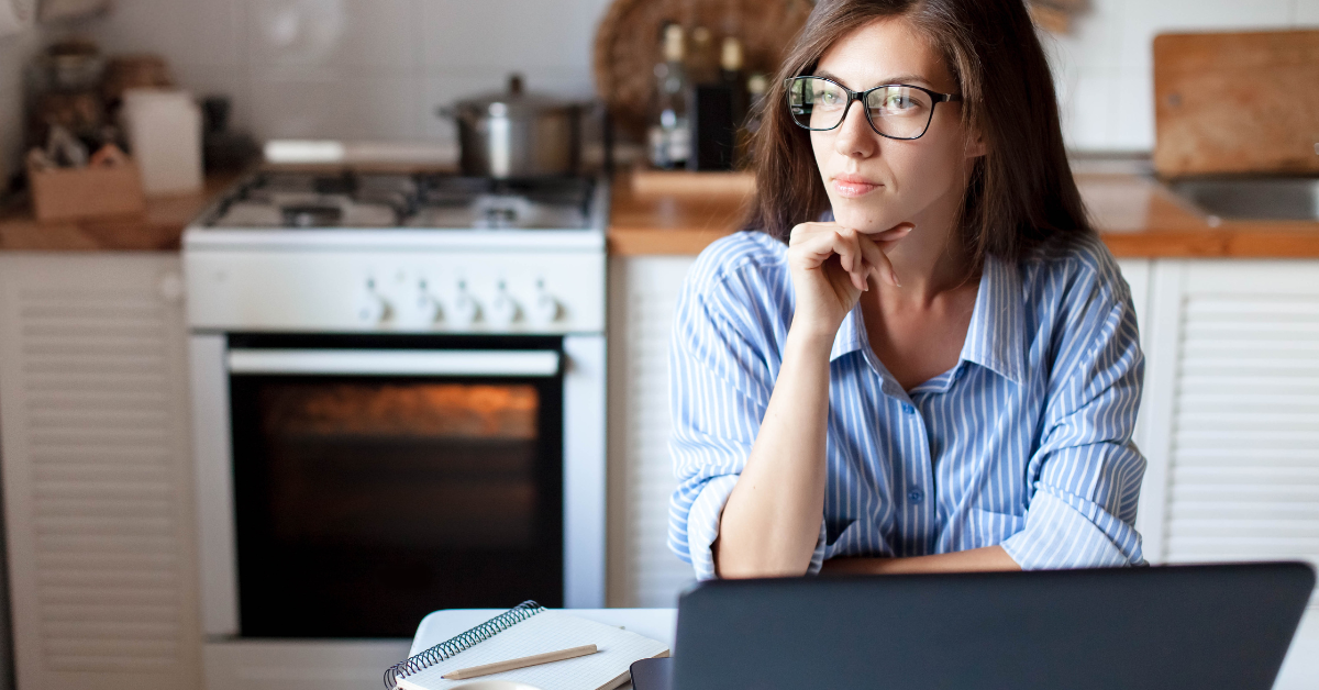 A woman sits at a kitchen table in front of a laptop, staring into the distance.