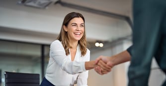 A smiling woman in a white blouse shakes hands with someone across a conference table in a bright office setting.