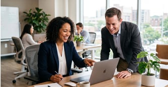 Two smiling business professionals, a seated woman in a navy blazer pointing at a laptop screen and a man in a gray suit jacket leaning in beside her, collaborate at a wooden desk in a bright, modern office.