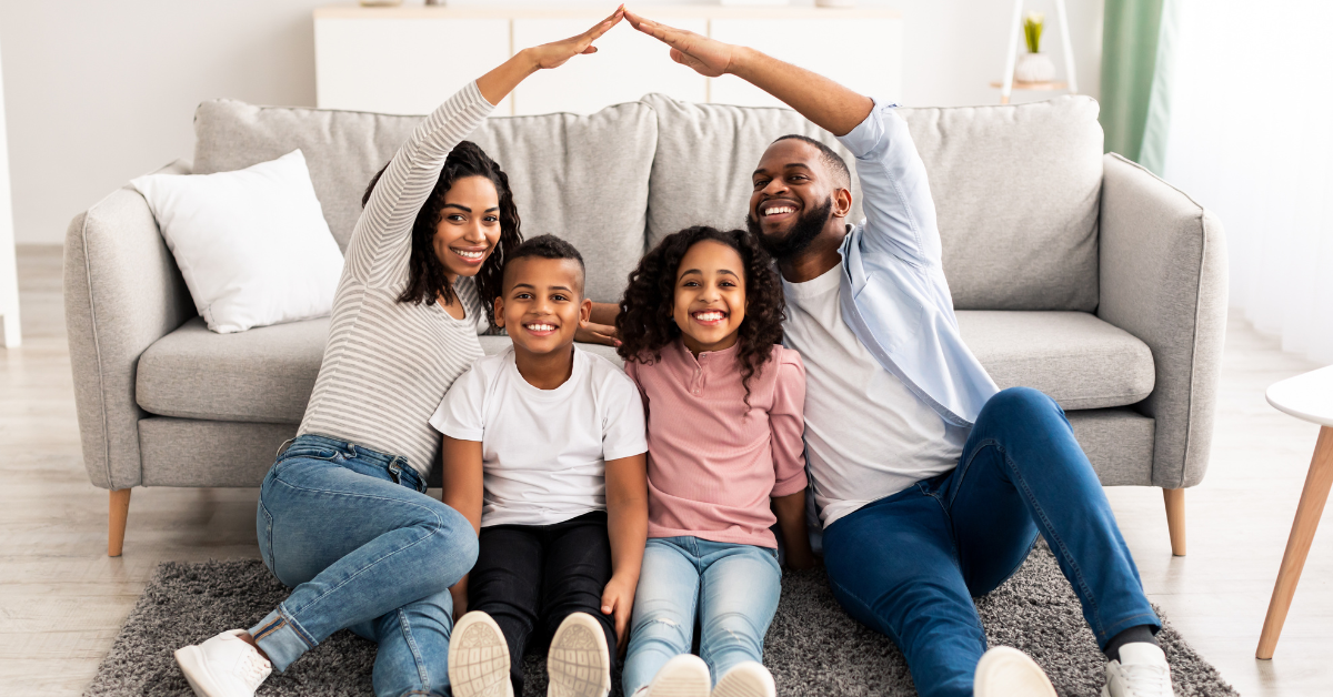 A smiling family of four sits in front of a couch.