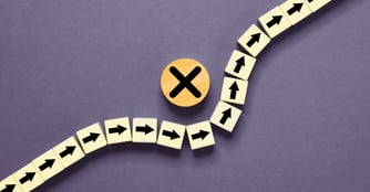 A row of light wooden tiles with black arrows points right along a path, then hits a wooden circle marked with a black X and is forced to detour upward in a new direction, symbolizing a blocked path or change in course.