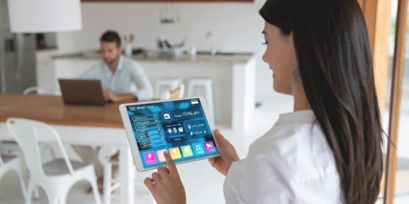 A woman taps the screen of a tablet in her kitchen while a man sits at a table with his laptop in the background.