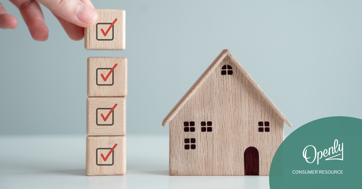 A person picks up a building block with a checkmark off of a stack of building blocks next to a wooden house. 