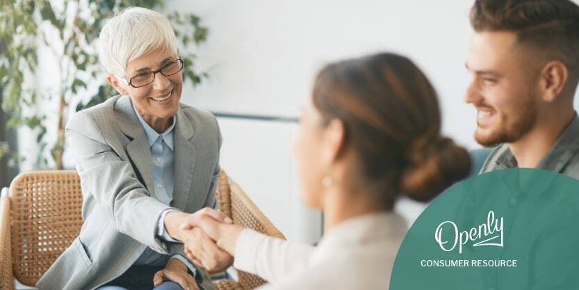 A business woman shakes hands with a woman while a man smiles in the background. 